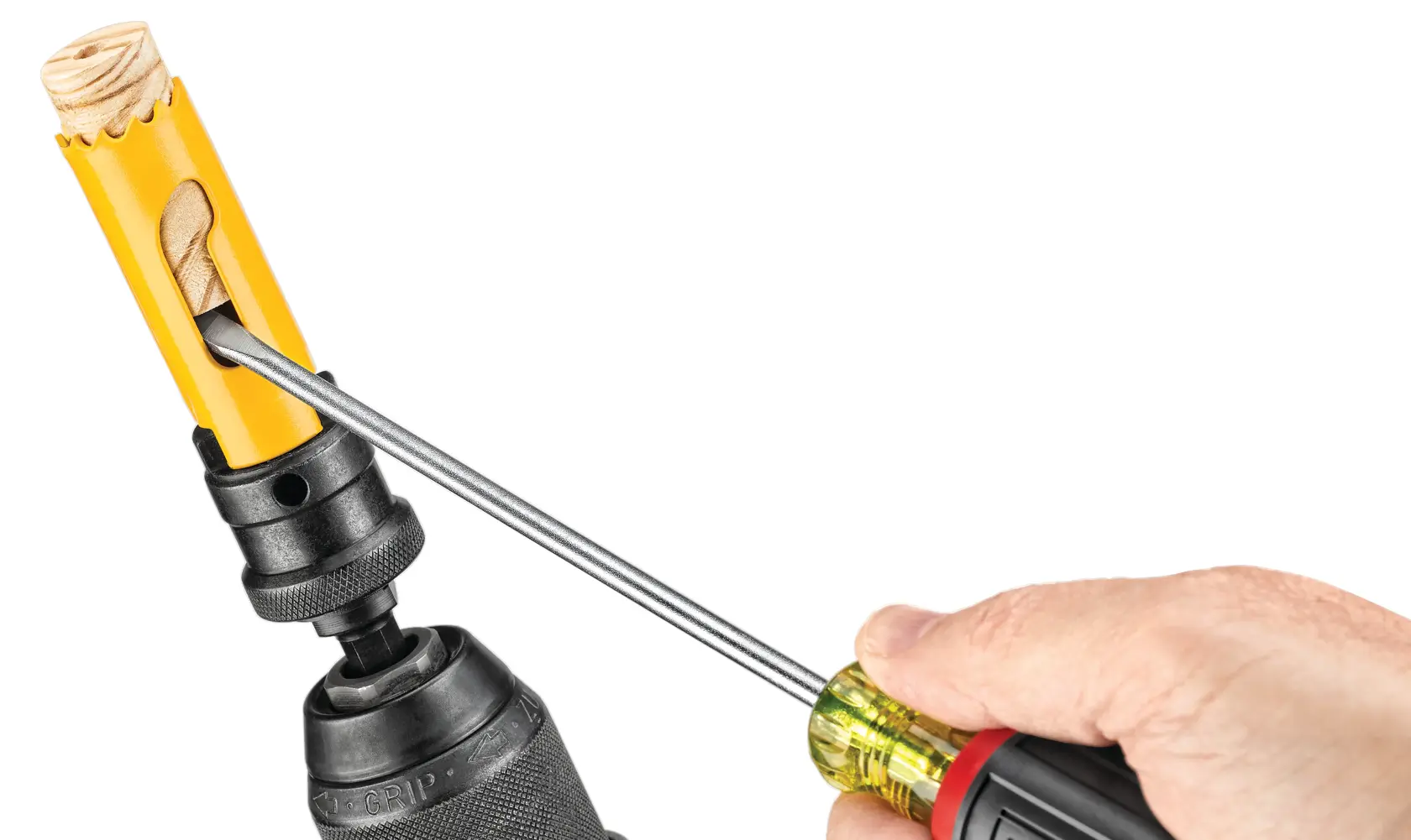A hand uses a screwdriver to remove wood debris from a yellow DEWALT hole saw attached to a drill.