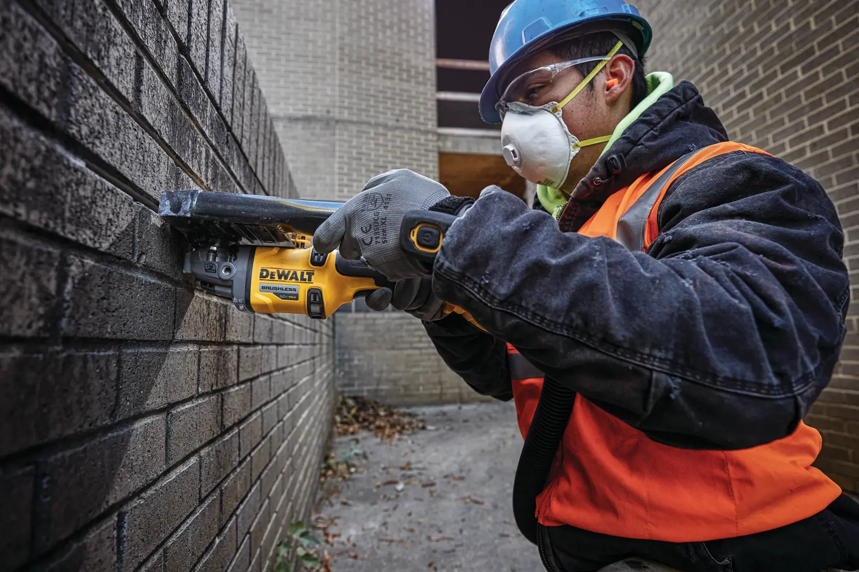 A person wearing protective gloves, a blue safety helmet, and an orange safety vest is using a yellow DEWALT power tool against a brick wall in an outdoor area.