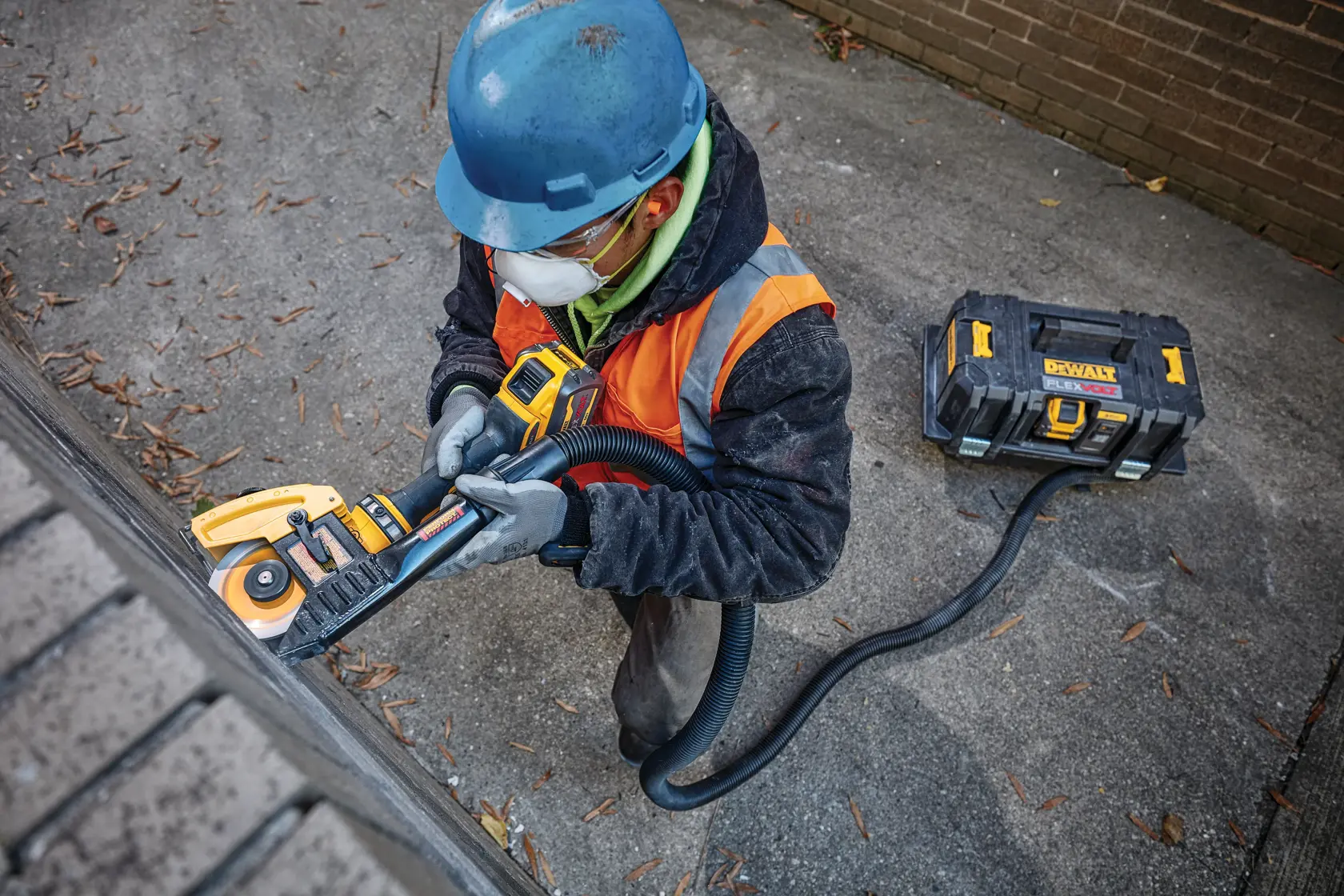 A worker wearing a blue hard hat, gloves, and orange safety vest operates a yellow DeWalt power tool connected to a portable vacuum system on an outdoor concrete surface.