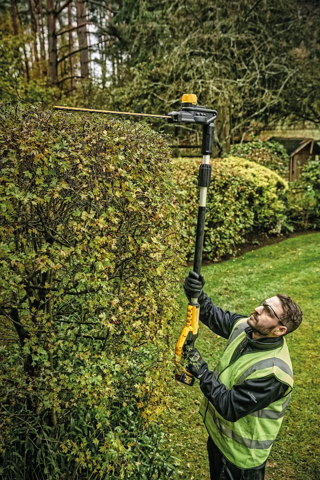 Overhead view of Pole Hedge Trimmer with battery being used by a person to cut through landscape outgrowth.