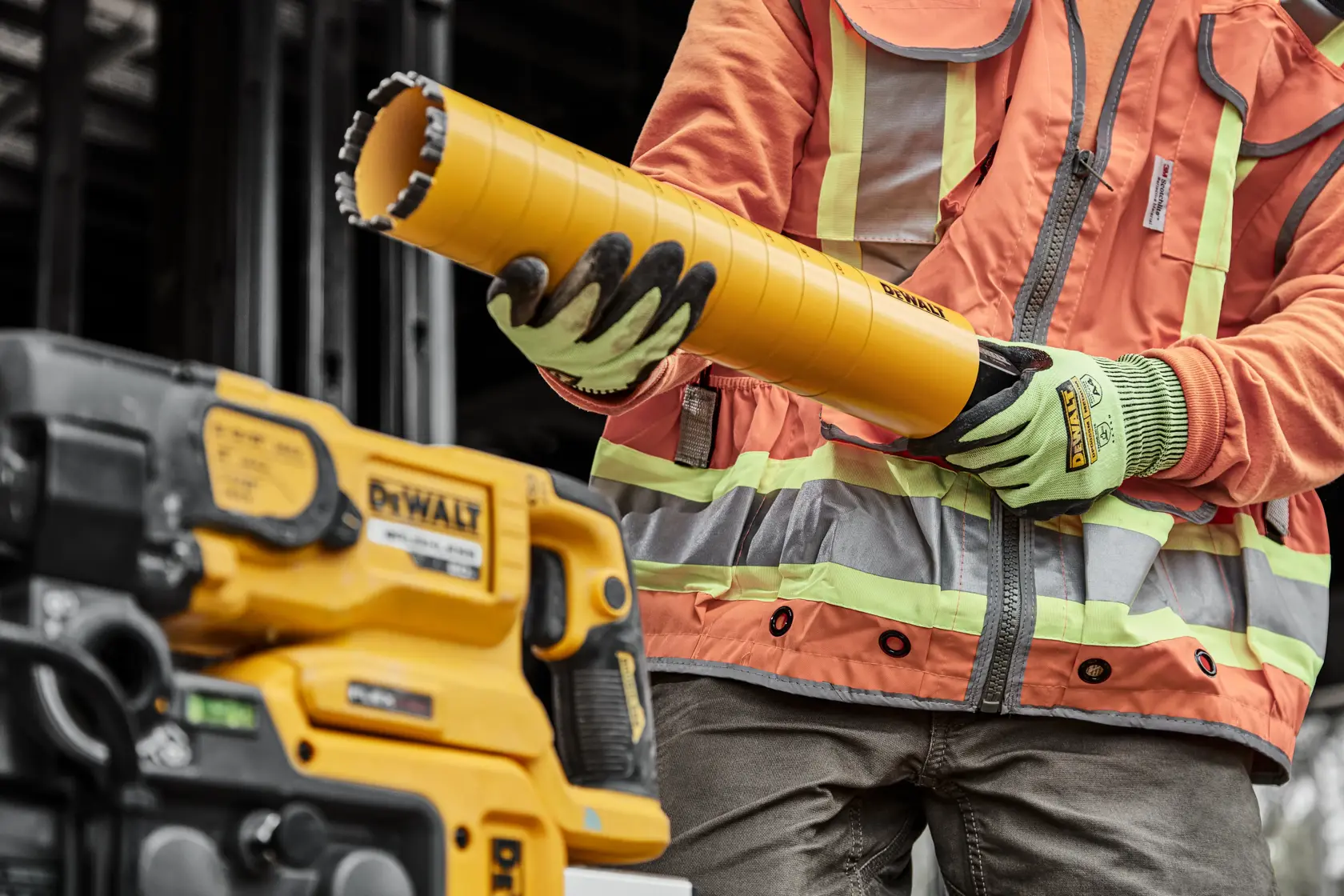 A person wearing a high-visibility orange and gray jacket and gloves is holding a large yellow DEWALT core drill bit next to a yellow DEWALT power tool at a construction site.