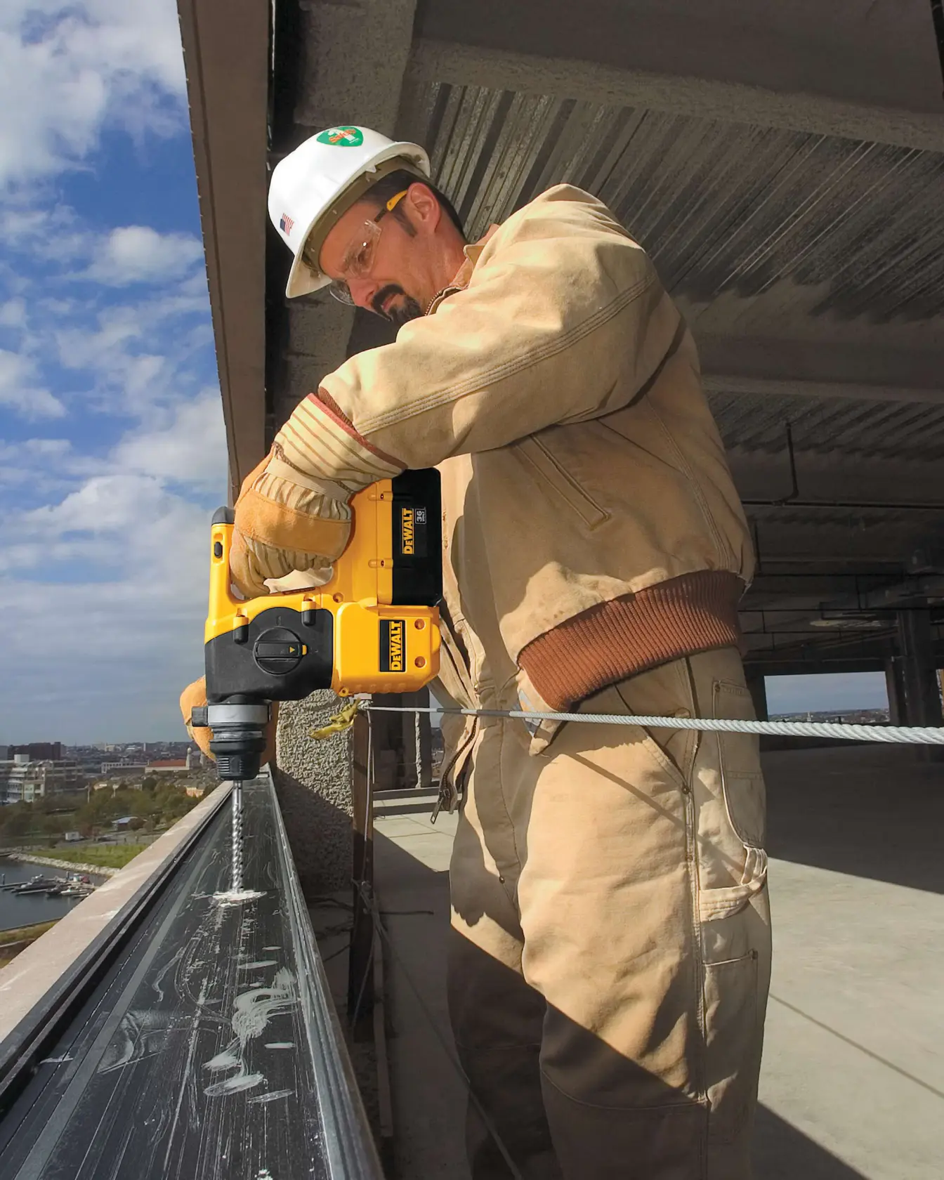 A person wearing protective clothing and gloves uses a yellow DEWALT power drill to drill into a metal beam at a construction site, with a safety harness attached and a city view in the background.