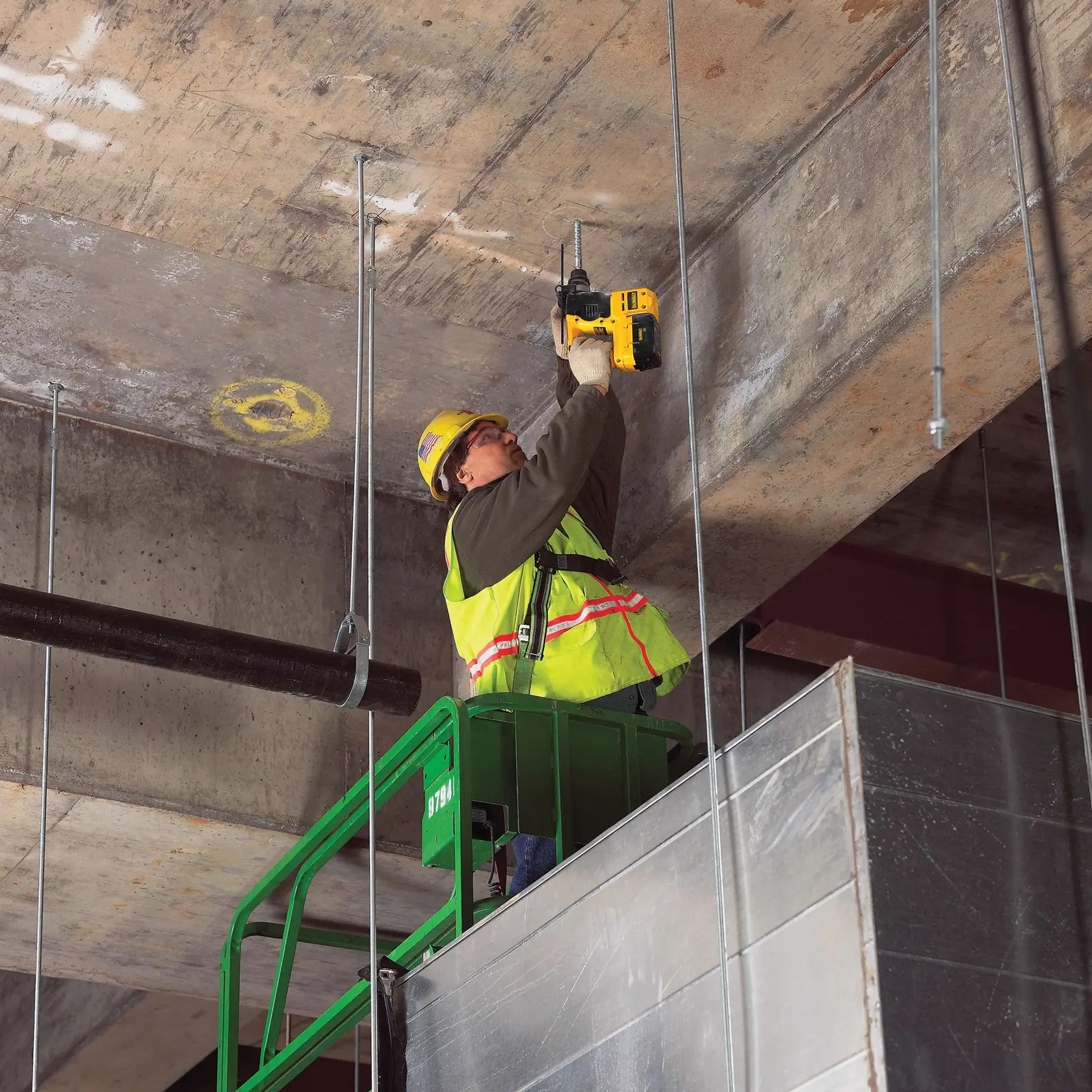 A construction worker using a DEWALT power tool to drill into a concrete ceiling while standing on a green lift. The worker is wearing a yellow safety vest and hard hat.