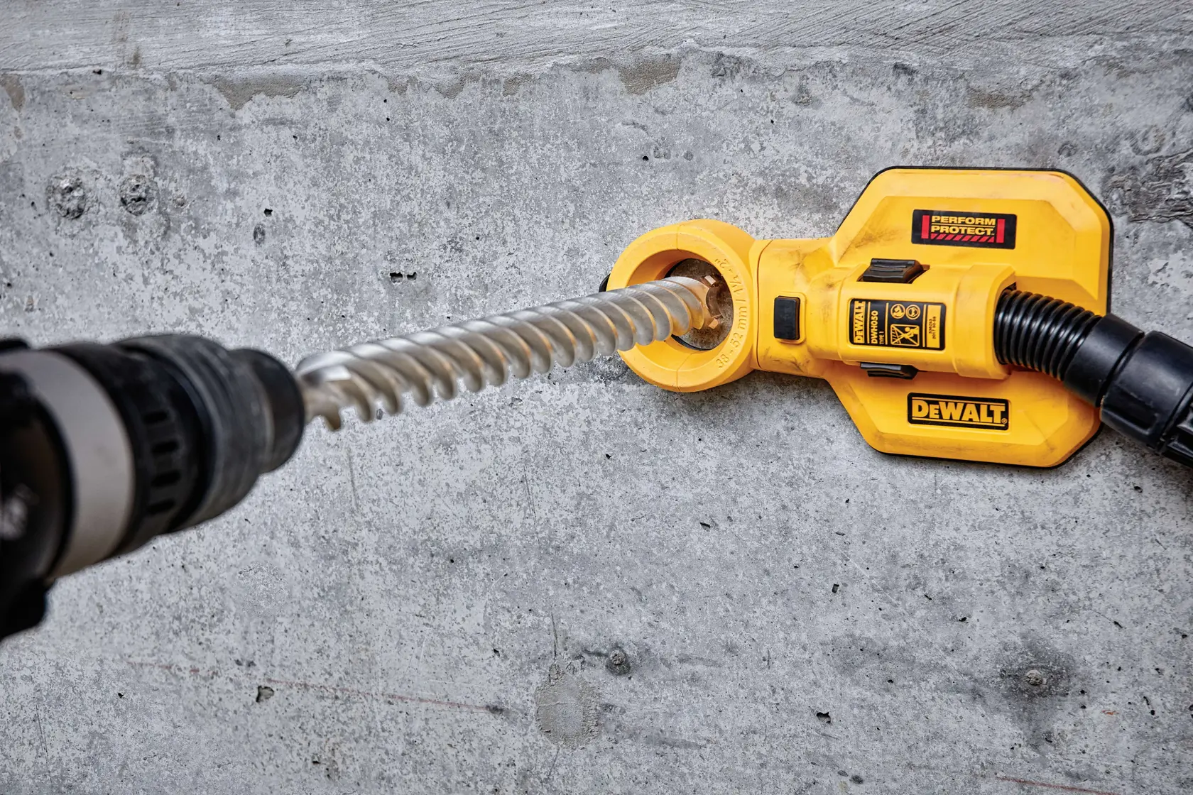 A DEWALT dust extraction attachment is connected to a power drill while drilling into a concrete wall. The yellow DEWALT device helps capture dust generated during the drilling process.