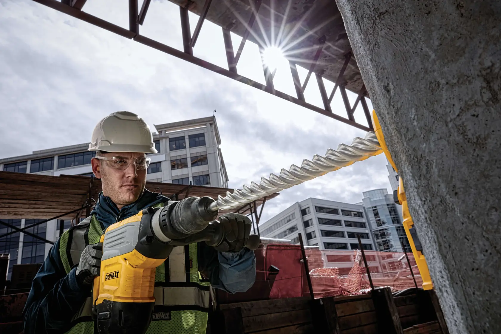 A construction worker using a DEWALT rotary hammer drill with a long spiral bit to drill into concrete at an outdoor construction site. The worker is wearing a hard hat and high-visibility vest. Modern buildings are visible in the background.