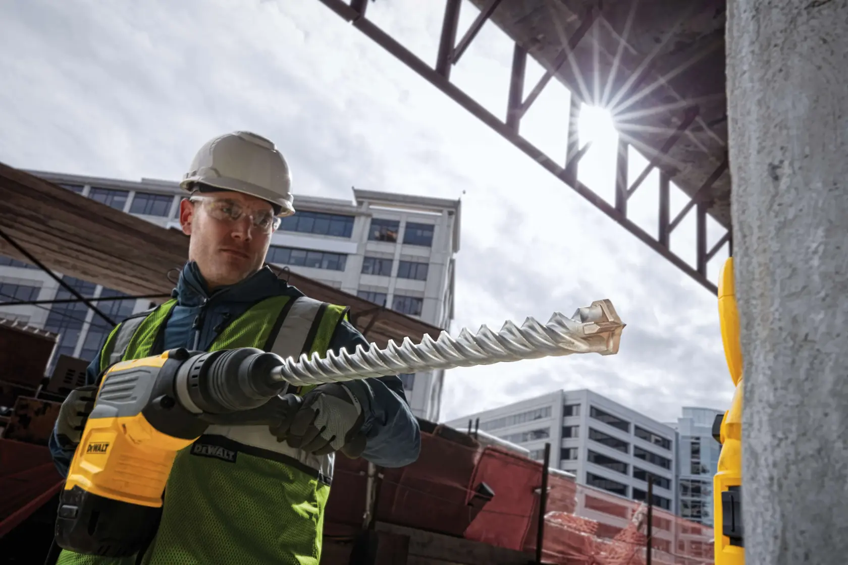 A construction worker wearing a safety vest and helmet operates a DEWALT rotary hammer drill with a large spiral drill bit at an outdoor building site. Modern buildings and construction structures are visible in the background.