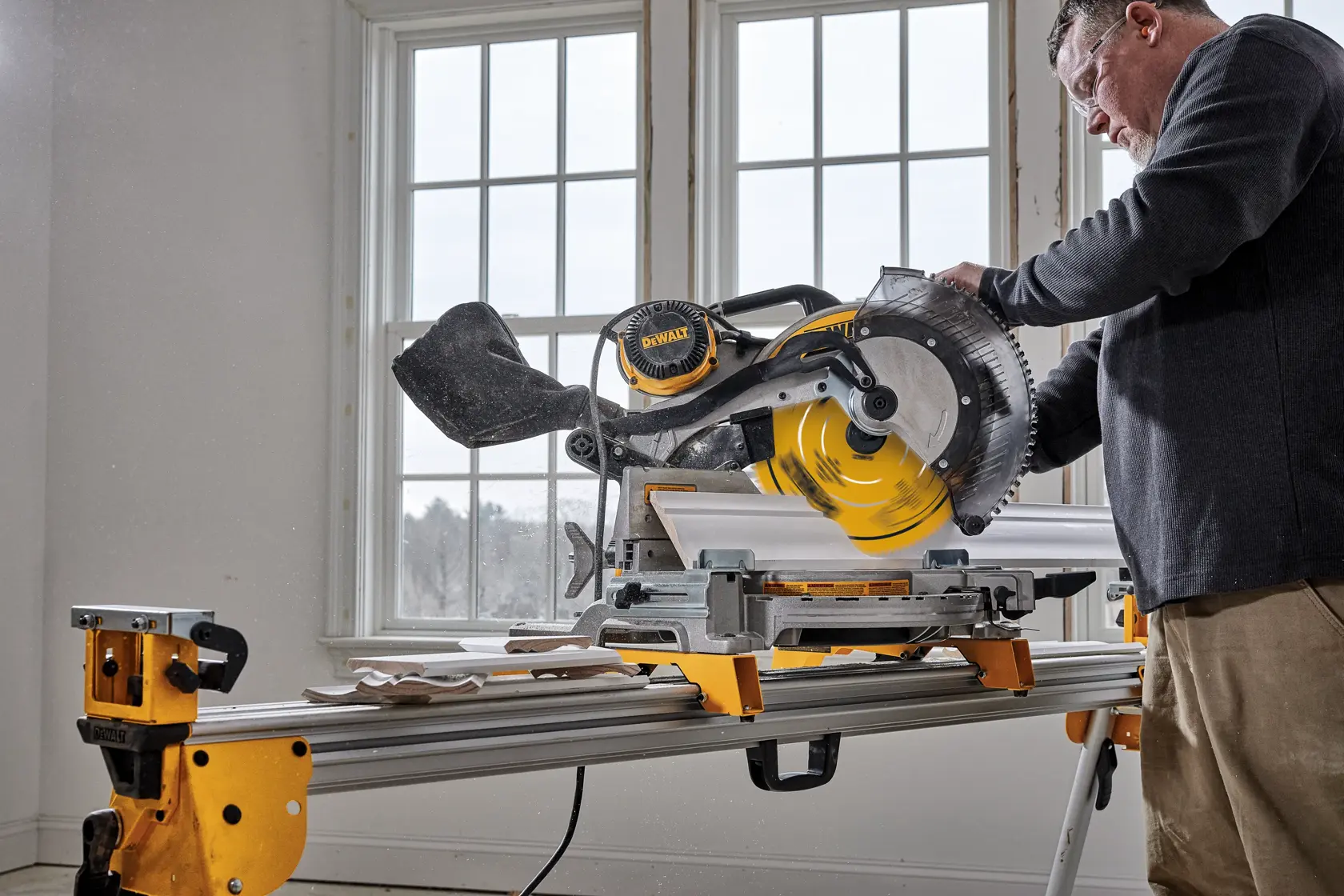 A person operating a DEWALT miter saw on a stand, cutting a piece of wood indoors near large windows.