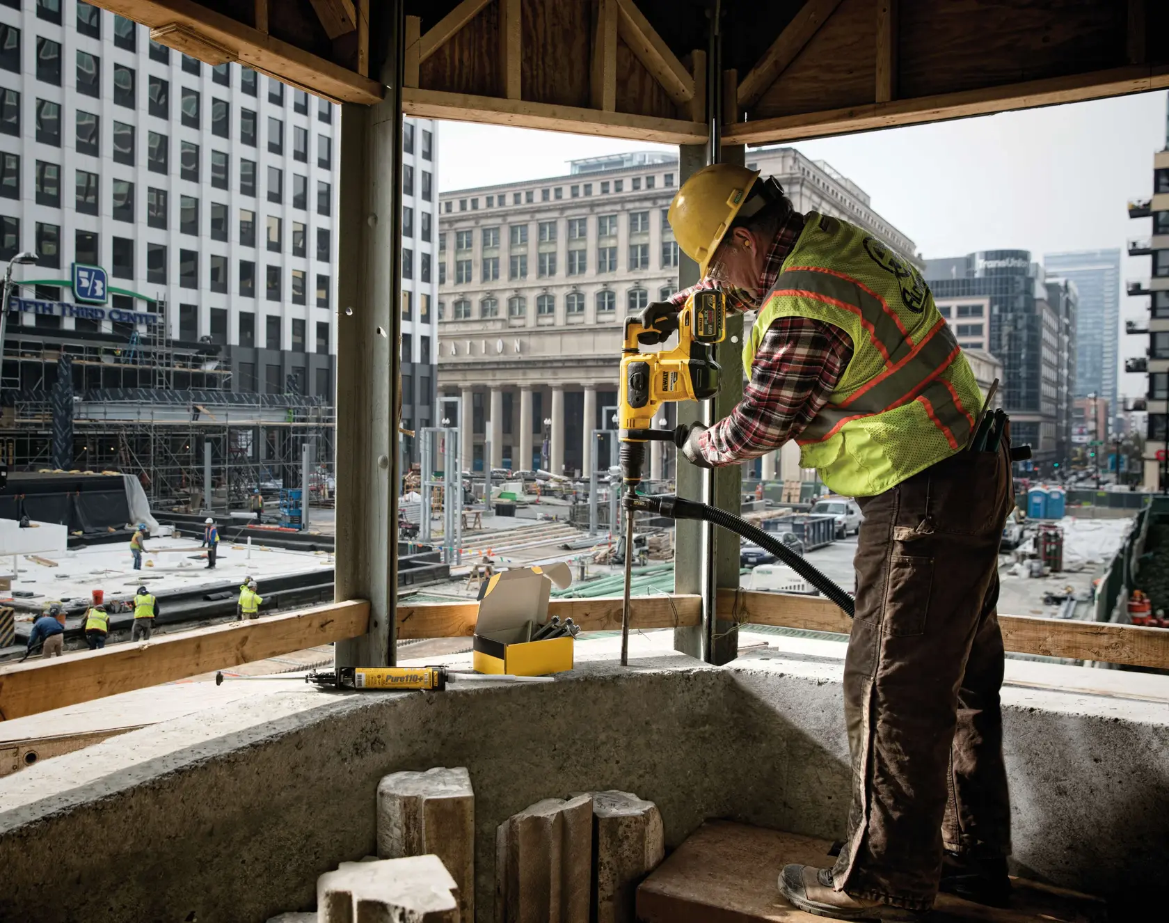 A construction worker wearing a yellow hard hat, safety vest, and protective glasses is drilling into a concrete surface with a DEWALT power tool at an urban construction site. The background shows several tall buildings and other workers operating on the site.