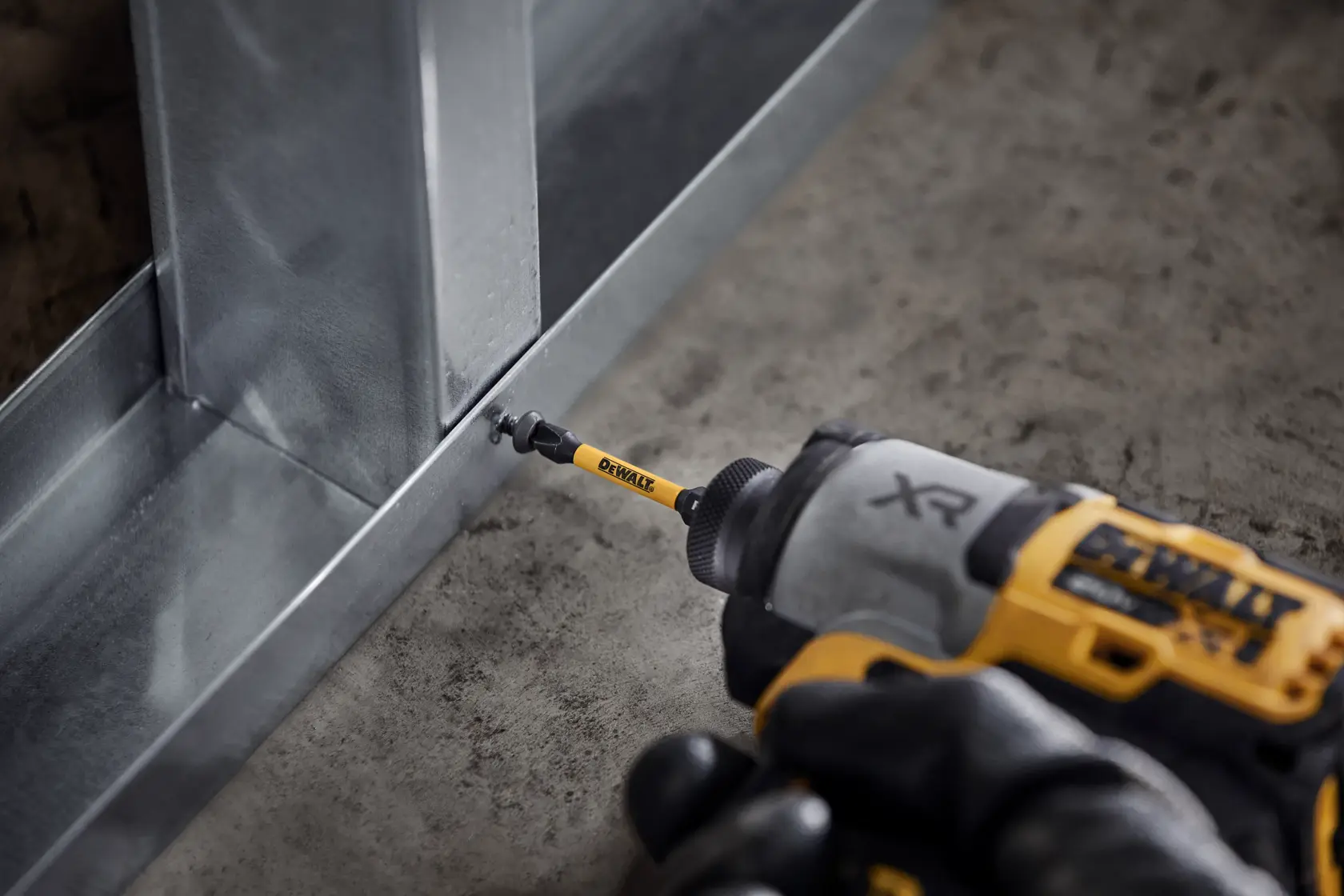 A DEWALT power drill with a yellow DEWALT bit is being used to fasten a screw into a metal frame on a concrete floor.