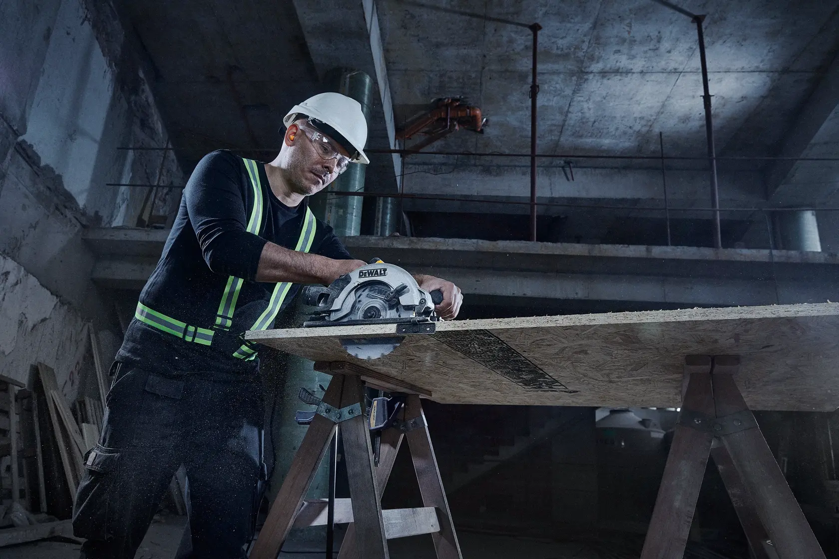 A person wearing a safety helmet and reflective vest is using a DEWALT circular saw to cut a wooden board supported by two sawhorses in a construction environment.