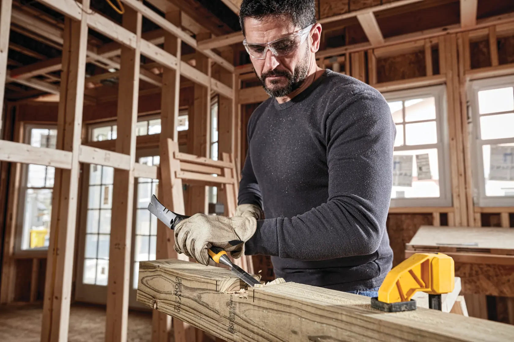 A person wearing gloves is using a DEWALT DWHT16861 tool to carve or work on a piece of wood clamped to a wooden structure in an unfinished room with visible framing and windows.