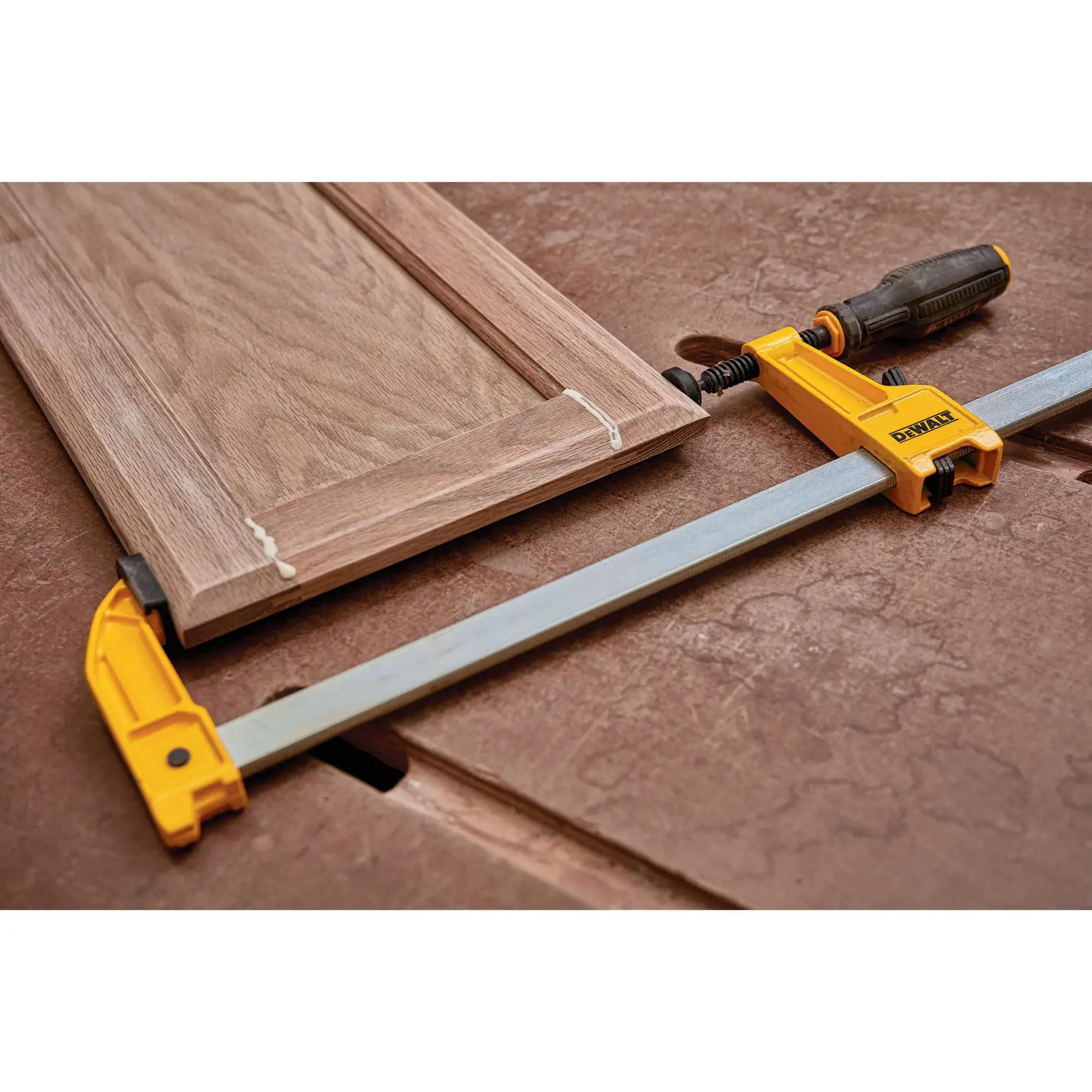A DEWALT bar clamp tool is being used to hold together a wooden cabinet door during the woodworking process. The clamp is yellow and black, with a sturdy metal bar across the wood piece, which appears to be glued at the joints.
