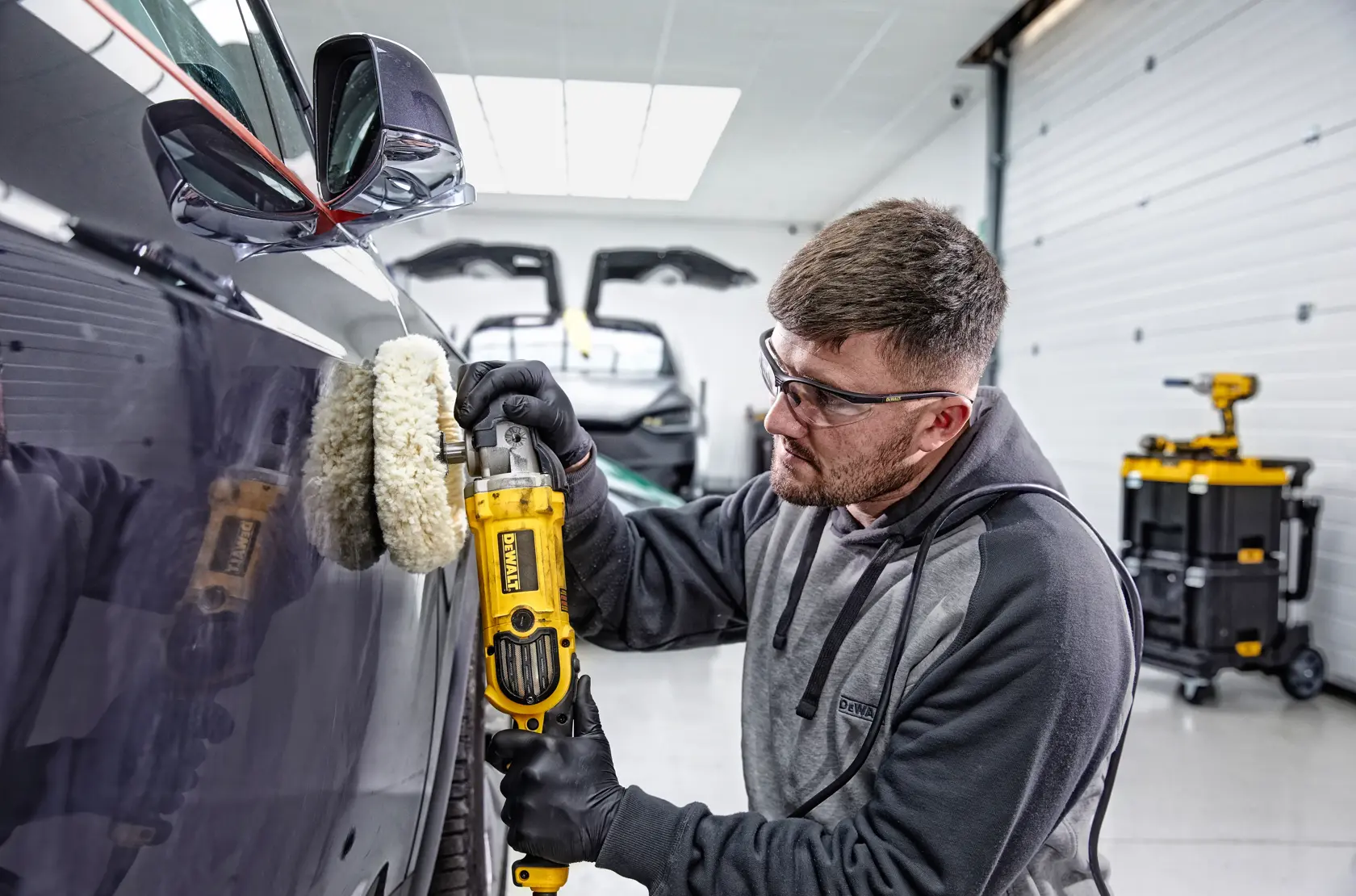 A person wearing gloves uses a DEWALT DWP849X polisher to buff the side of a dark-colored car in a garage. The person's face is blurred, and there are tools and equipment visible in the background.