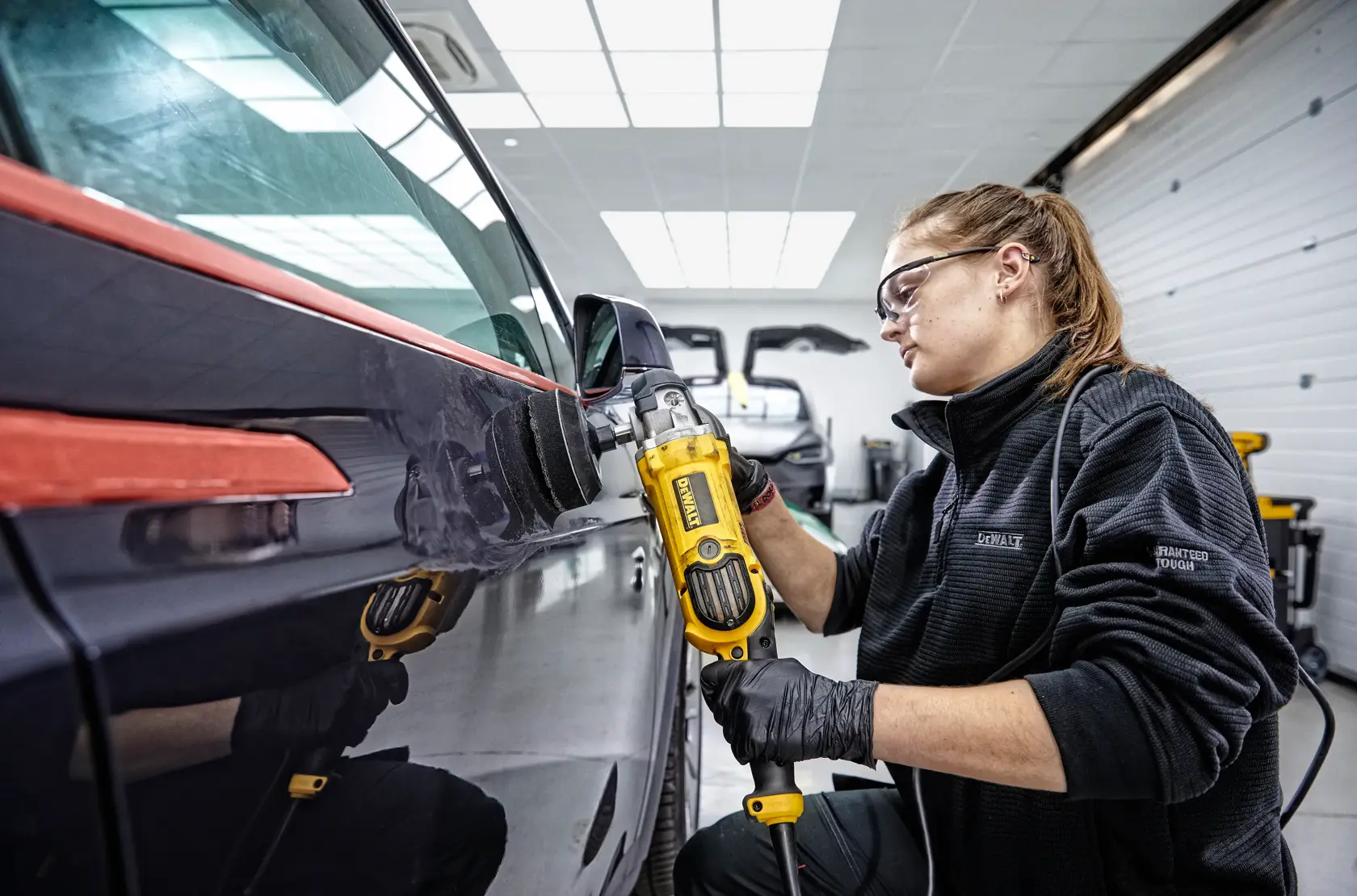Person using a DEWALT DWP849X polisher on a car surface in a workshop. The individual is wearing black gloves and a dark jacket, and the setting is a brightly lit garage.