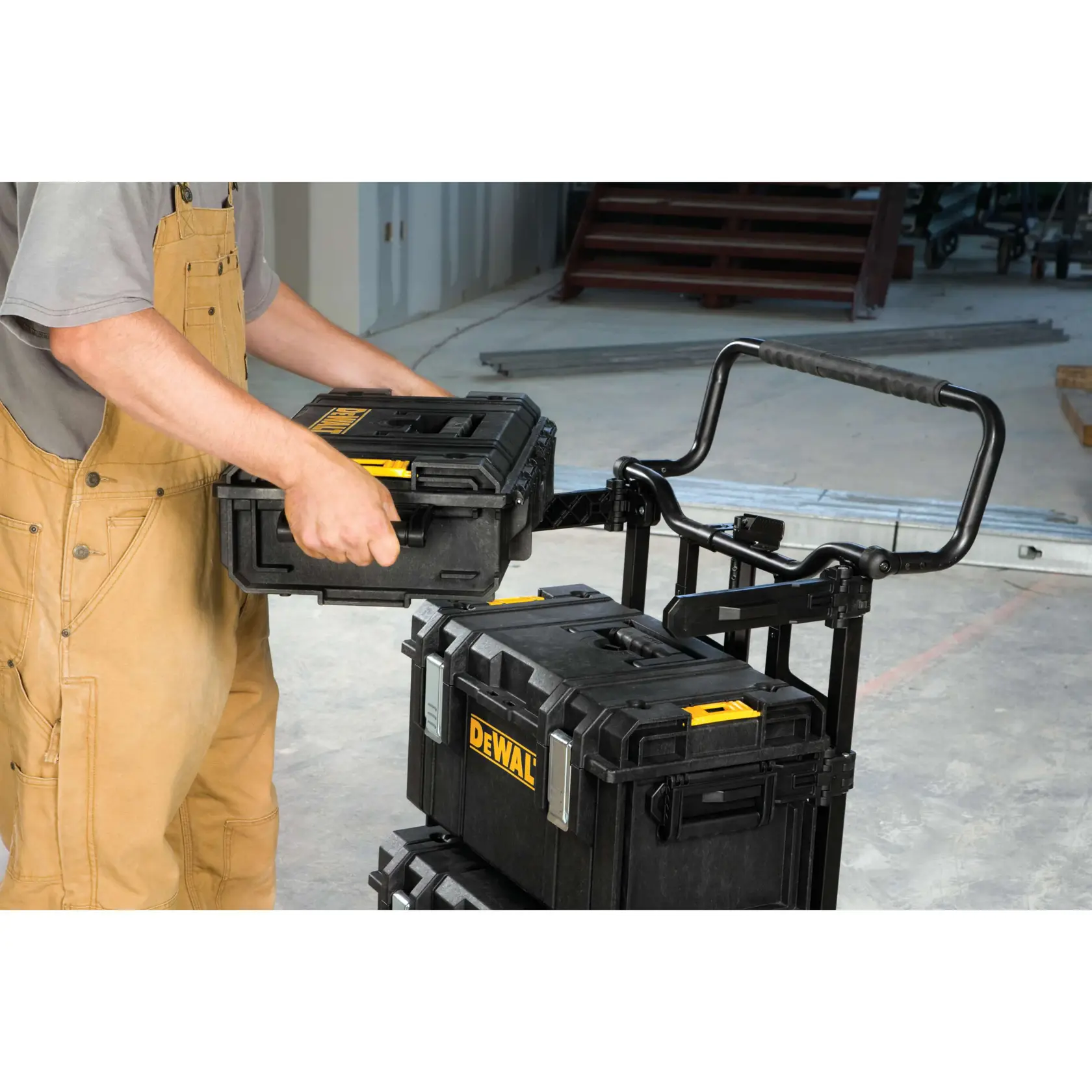 A person wearing brown overalls is stacking DEWALT black toolboxes on a rolling cart inside a workshop or construction area.