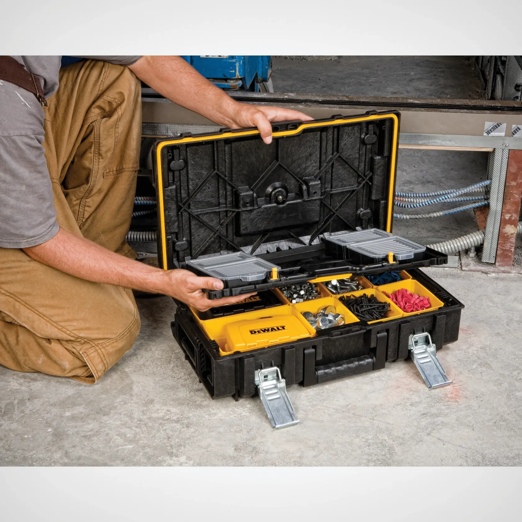 A person kneeling on a concrete floor, opening a black and yellow DEWALT organizer toolbox filled with various hardware items such as screws, bolts, and fasteners. The toolbox features multiple compartments for sorting tools and accessories.