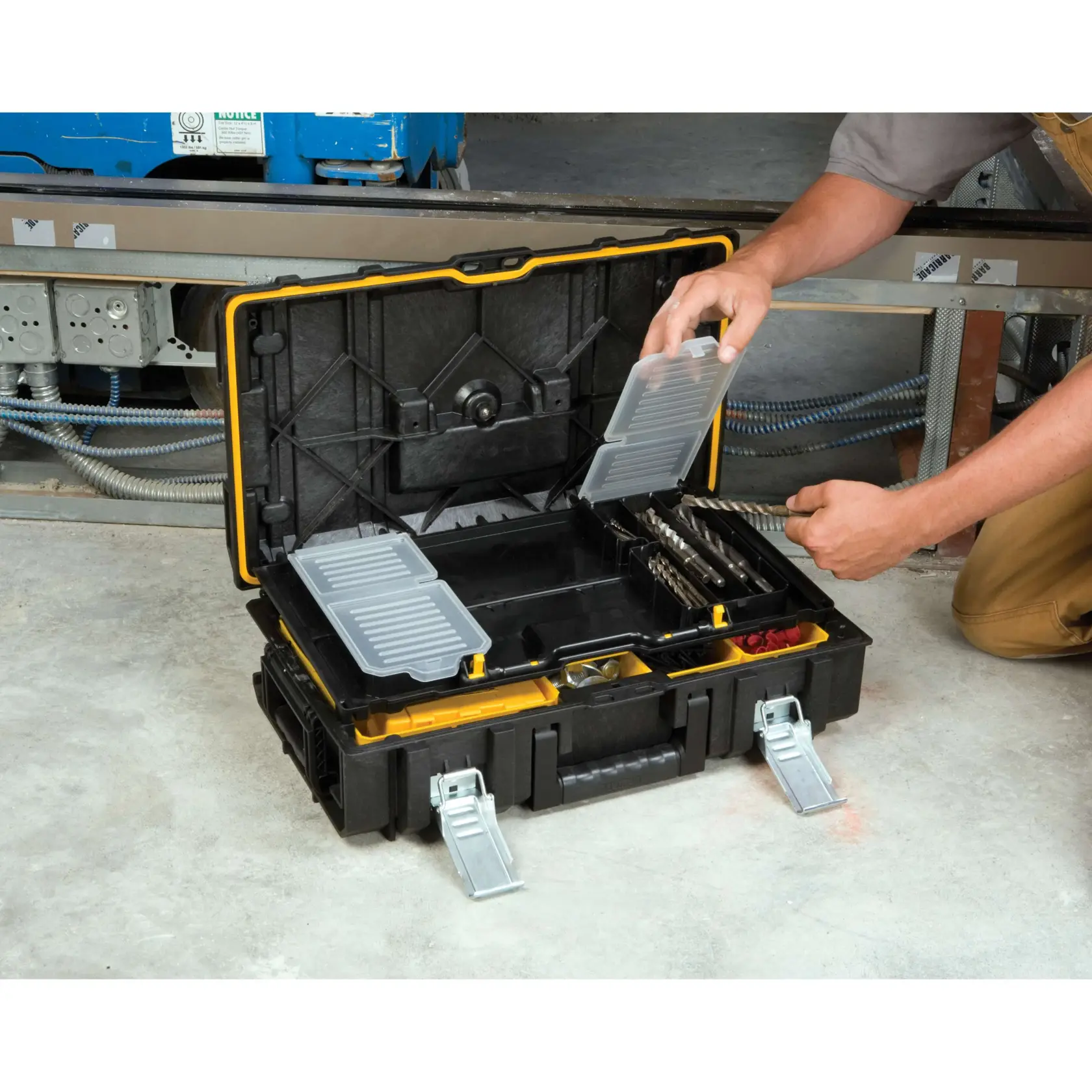 A person is organizing drill bits in a DEWALT tool storage box with open compartments and metal latches, positioned on a concrete floor in a workshop setting.