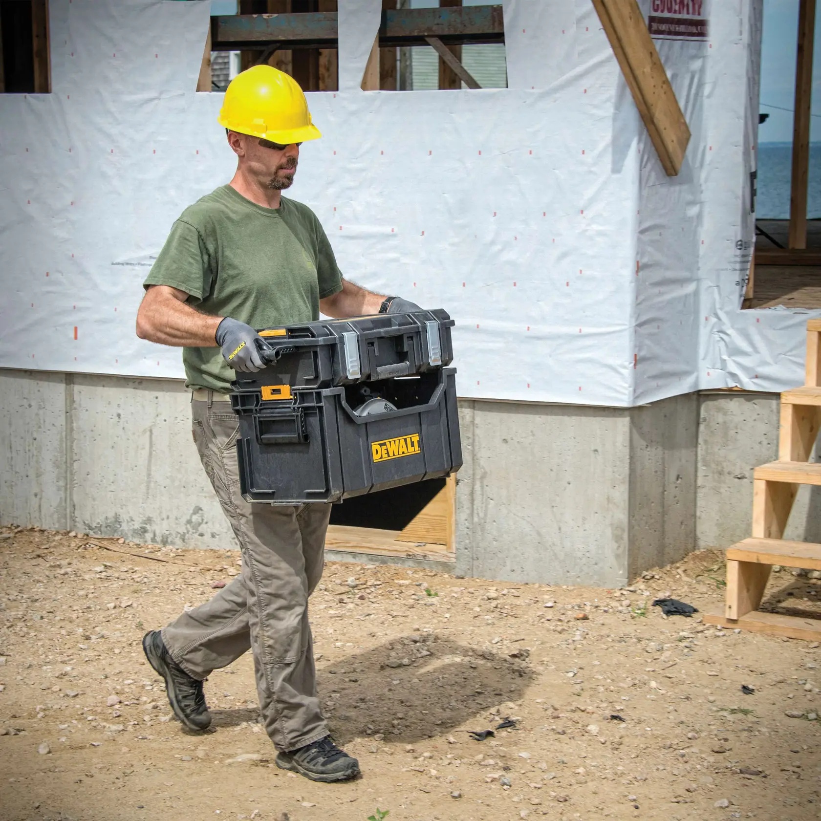 A person wearing a yellow hard hat and gloves is carrying a DEWALT DWST08205 black toolbox outside at a construction site.