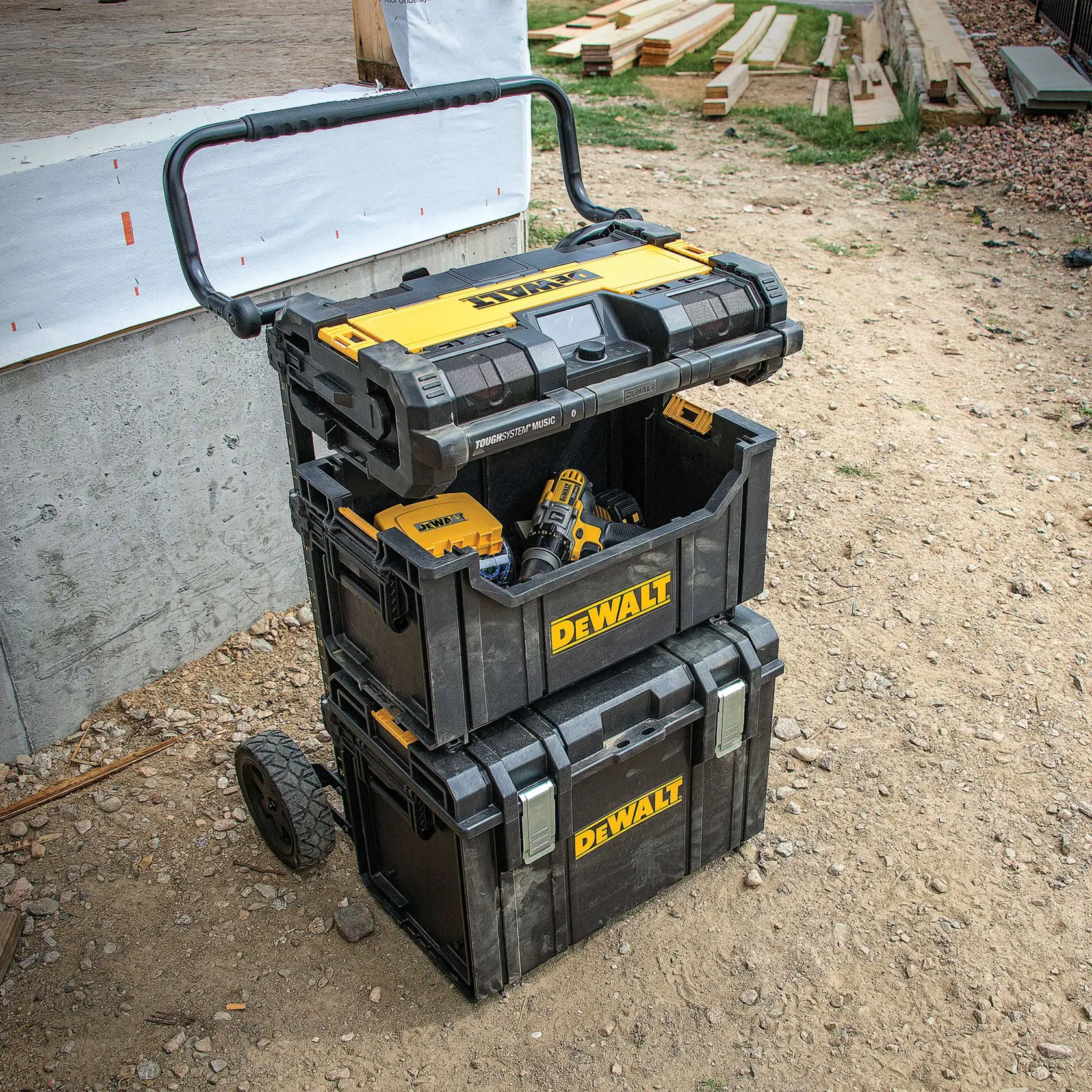 A stackable DEWALT mobile tool storage system with three black and yellow toolboxes on rugged wheels and a handle, shown outdoors at a construction site. The middle box contains various tools, including a cordless drill.