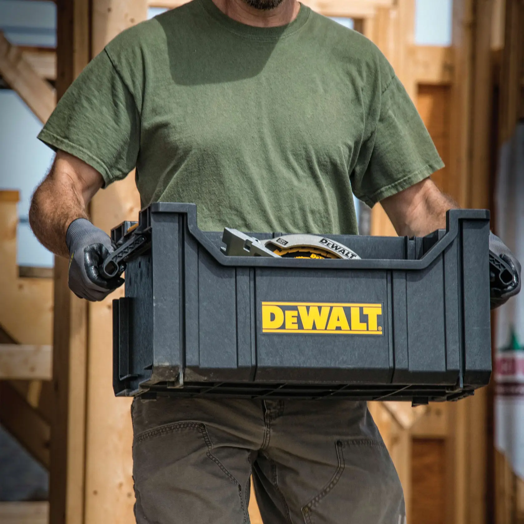 A person wearing a green shirt and gloves carrying a black DEWALT branded toolbox with tools inside at a construction site.